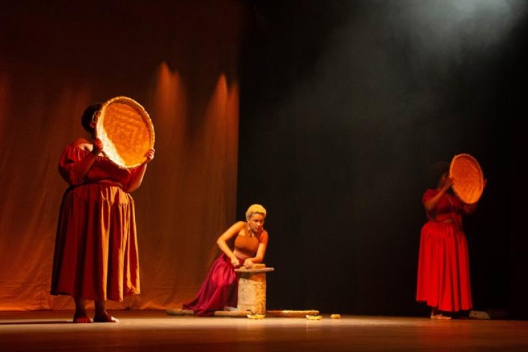 Teatro Marília recebe a 10ª edição da Mostratu com cenas curtas do Teatro Universitário da UFMG Cena teatral com três mulheres em um palco. À esquerda, em primeiro plano, uma mulher de pele negra veste um vestido longo e rodado na cor terracota. Ela está em pé, descalça, segurando uma peneira à frente do próprio rosto. Ao centro, uma jovem mulher de cabelos loiros curtos e pele clara está sentada no chão. Ela veste um top marrom e uma saia longa vinho, apoiando os braços em um pequeno cepo. À direita, em segundo plano, uma terceira mulher também encobre o rosto com uma peneira.