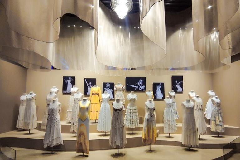 Exposição “Clara Nunes – Eu Sou a Tal Mineira” se despede do público após temporada de sucesso no Mumo "Fotografia da exposição 'Clara Nunes - Eu Sou a Tal Mineira'. Em uma sala com iluminação suave e teto decorado com tecidos ondulados, vários manequins exibem figurinos originais da cantora. A maioria são vestidos longos em tons brancos e cremes, com detalhes em rendas e franjas, além de um vestido amarelo ao centro-esquerda. Ao fundo, quadros em preto e branco mostram fotos de Clara Nunes no palco."