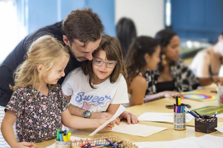 Museus municipais estão com programação diversificada neste mês de setembro Em uma sala de atividades, crianças e adultos participam de uma oficina criativa ao redor de uma mesa com papéis, desenhos, canetas e lápis coloridos. Em primeiro plano, um homem acompanha duas meninas durante a atividade, enquanto outros participantes trabalham ao fundo.