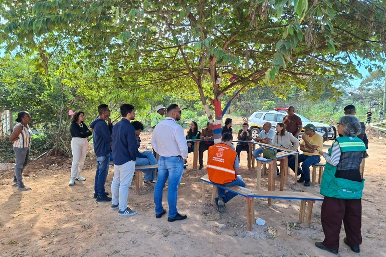 PBH e moradores promovem mutirão de limpeza e conscientização ambiental no Novo Aarão Reis Serviores da prefeitura, usando uniforme laranja e verde, conversando com pessoas sentadas em bancos colocados ao lado de árvores e viatura da guarda municipal