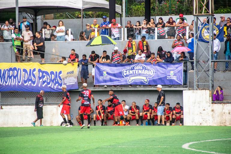  Fotografia de uma partida da Copa Centenário de Futebol Amador. Em primeiro plano, jogadores de uniforme preto e vermelho estão próximos ao banco de reservas, ao lado do campo de grama sintética.