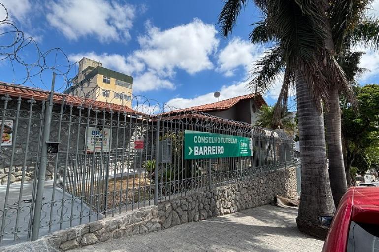 Conselho Tutelar Regional Barreiro funciona em novo endereço para mais qualidade do atendimento à população Fotografia externa, durante o dia, da fachada de um imóvel onde funciona o Conselho Tutelar Barreiro. O prédio é cercado por um muro baixo de pedras e um portão de grades metálicas, sobre o qual há várias voltas de arame farpado. Ao centro da imagem, preso às grades, há uma grande placa verde com a inscrição “CONSELHO TUTELAR BARREIRO”, acompanhada de uma seta apontando para a direita e da identidade visual da Prefeitura de Belo Horizonte.