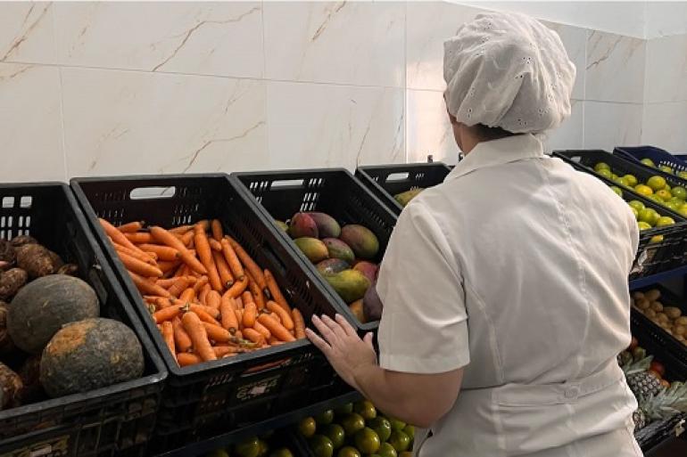 Nutricionistas garantem a qualidade da alimentação nas unidades da Política de Segurança Alimentar e Nutricional de BH Imagem mostra uma mulher de costa vestida com uniforme e toca brancos. Ela está diante de uma banca com abóboras, cenouras e outros legumes.