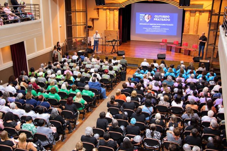 Seminário do Outubro Prateado marca a abertura oficial do Mês de Valorização da Pessoa Idosa Imagem de auditório lotado com palestrante à frente, no palco, falando sobre o tema Outubro Prateado.