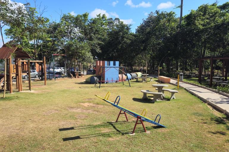 A imagem mostra um amplo gramado, onde estão instalados brinquedos infantis como gangorra,, escorregador, casinha de madeira, além de mesas de concreto com bancos, uma pista de caminhada e um pergolado sob árvores do Parque do Brejinho.