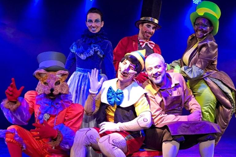 Uma fotografia colorida em plano médio de seis atores fantasiados, posando sorridentes em um palco de teatro iluminado por luzes azuis.