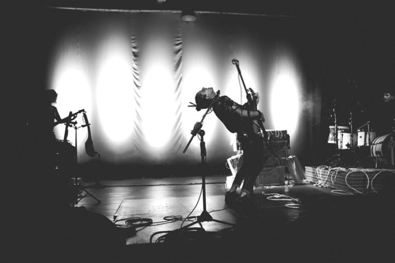 Show no Teatro Marília promove encontro entre sonoridades mineiras e afro-latinas Fotografia em preto e branco de uma apresentação musical no palco. No centro, Ricardo Ulpiano canta inclinando o corpo para trás diante do microfone enquanto toca guitarra, sob a iluminação de refletores ao fundo.