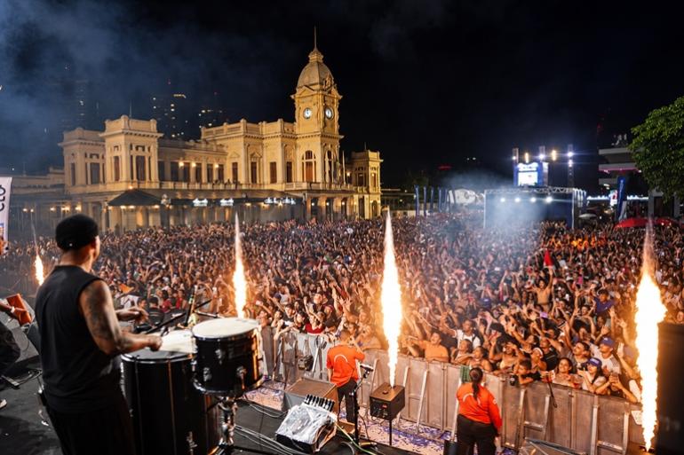 Prazo para agendar eventos de rua em BH no 1º semestre de 2027 termina em 31 de outubro Um músico toca tambores no palco diante de uma grande multidão animada, com braços erguidos, na Praça da Estação, em Belo Horizonte. Jatos de fogo iluminam o ambiente, enquanto, ao fundo, destaca-se o prédio histórico do Museu de Artes e Ofícios, com sua torre de relógio.