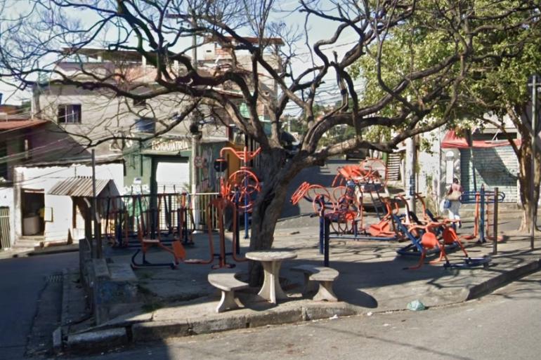 Imagem mostra uma praça circular com brinquedos, mesa, bancos e uma árvore. Hácasas em volta.
