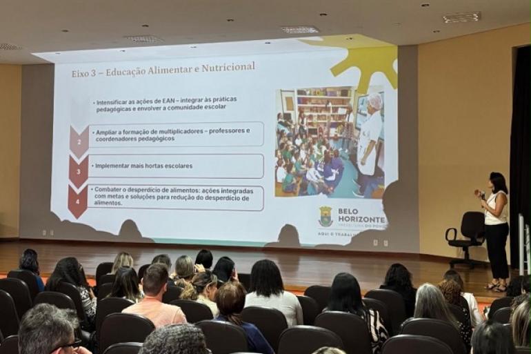 Imagem mostra um auditório com pessoas de costas e uma mulher no palco apresentando um power point com informações sobre alimentação nutricional.
