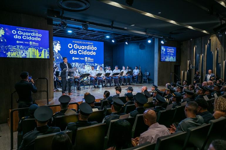 Secretário Márcio lobato discursa na solenidade de entrega do pr~emio, tendo ao fundo um banner com uma lausão ao nome da premiação . No palco há também todos os membros da banda e em frente, de costas, os comvidados.