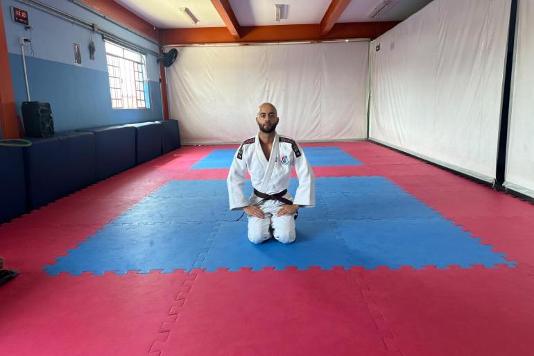 Em uma sala de treinamento de artes marciais, o professor Igor Oliveira posa, ajoelhado, em um tatame azul e vermelho. Igor usa quimono branco. Ao fundo e ao lado, há paredes brancas e azuis.m.