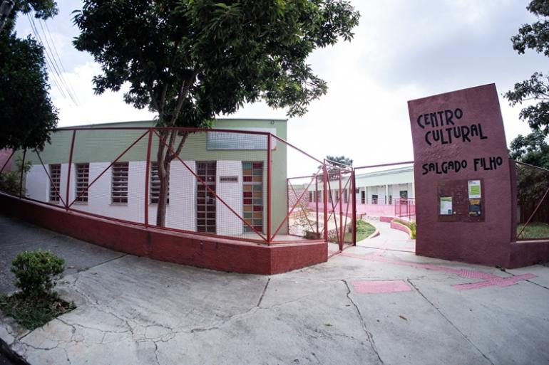 Centros culturais das regionais Oeste e Noroeste comemoram o Mês do Patrimônio com programação diversificada Fachada do Centro Cultural Salgado Filho. A imagem destaca o totem de identificação na cor vinho com o nome do centro esculpido em preto, e a grade metálica vermelha que cerca a área externa. Ao fundo, o prédio moderno com paredes verdes claras e janelas com detalhes de vidros coloridos (vermelho, amarelo, azul).