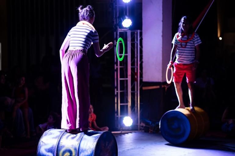Fotografia num ambiente cénico e iluminado que mostra dois artistas circenses a realizarem uma performance de equilíbrio.Em primeiro plano, à esquerda e de costas, uma artista equilibra-se num barril metálico deitado, vestindo camisola de riscas e calças cor-de-rosa. No ar, entre os dois, flutua um arco verde-neão. À direita, outro artista equilibra-se noutro barril com calções vermelhos e camisola às riscas, segurando um arco laranja. O fundo é escuro, com refletores de iluminação e o público visível à vol