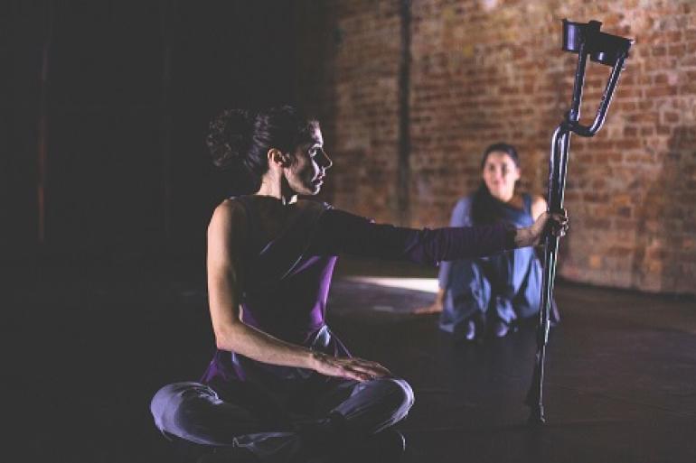 Festival Acessa BH transforma o Teatro Raul Belém Machado em palco da inclusão A imagem mostra, em primeiro plano, uma mulher de pele clara e cabelo escuro preso, que está sentada de pernas cruzadas sobre o chão escuro de um palco. Ela veste uma blusa roxa e calça cinza. Seu corpo se encontra voltado de perfil para a direita e a cabeça está virada em direção a uma muleta que ela segura com o braço direito estendido, mantendo o objeto equilibrado na vertical à sua frente. Ao fundo, ligeiramente desfocada, outra mulher senta-se no chão vestindo uma roupa azul-escura, observando a cena.
