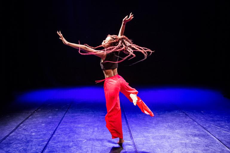 Centro de Referência da Dança oferece ocupação gratuita para artistas e coletivos Mulher dançando no palco, sendo iluminada por luz azulada, usando calça vermelha e blusa preta