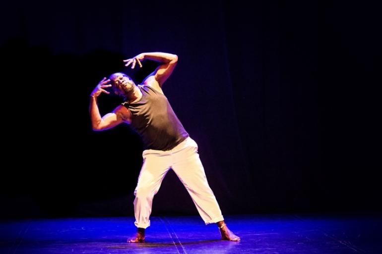PBH fortalece a pesquisa em dança com nova edição do Edital Corpo-Território Homem negro dança descalço no centro de um palco escuro, sob iluminação amarelada e chão azul. Ele veste regata preta e calça branca. Inclina o tronco para a direita com as pernas abertas, joelhos dobrados e as mãos espalmadas ao redor da cabeça, em expressão concentrada.