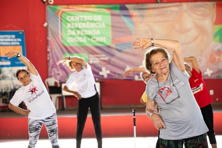  Grupo de mulheres idosas participa de uma atividade de alongamento. Em primeiro plano, uma mulher de cabelos curtos usa camiseta cinza e inclina o corpo para o lado, mantendo um dos braços levantado sobre a cabeça. Atrás dela, outras participantes repetem o movimento. Ao fundo, um grande painel colorido traz a inscrição “Centro de Referência da Pessoa Idosa – CRPI”.