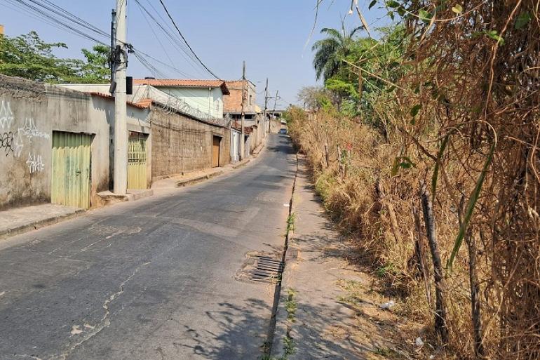 Fotografia de uma rua residencial em área urbana. A via é asfaltada, estreita e segue em aclive. À esquerda, há casas simples, muros e portões, além de um poste de concreto com vários fios de energia. À direita, há uma calçada estreita e um terreno com vegetação alta, seca e bastante densa. No centro da rua, mais ao fundo, há um carro estacionado. O céu está claro e o dia aparenta estar ensolarado.