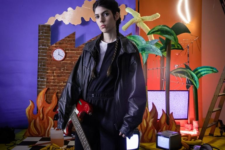“Sara Não Tem Nome” apresenta show e revela bastidores de novo videoclipe no Teatro Marília A fotografia mostra uma pessoa jovem de pele clara, cabelos escuros presos em duas tranças longas e expressão séria, posicionada no centro da cena e olhando diretamente para a câmera. Ela segura uma guitarra elétrica vermelha com o braço inclinado para a frente. Veste uma jaqueta de couro preta oversized por cima de uma blusa preta e branca com zíper e calça escura. O cenário ao fundo é vibrante, lúdico e cênico, dividido ao meio por cores contrastantes.
