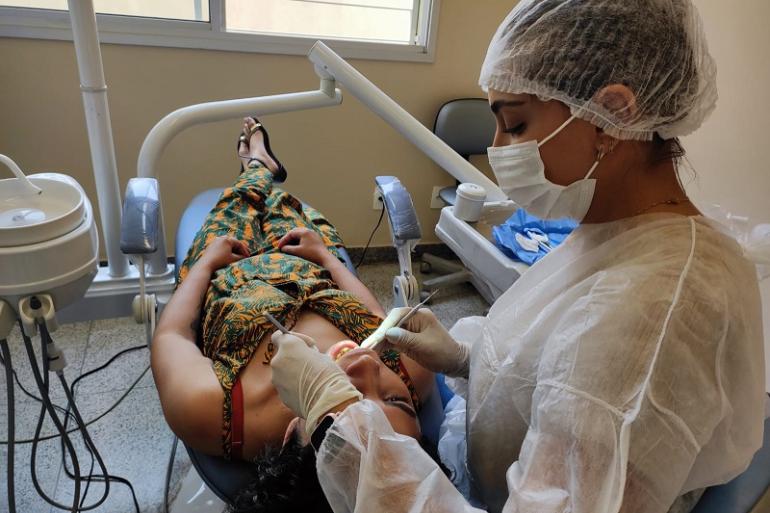 PBH promove primeiro Fórum de Saúde Bucal da Região Metropolitana para ampliar o cuidado com a população A foto mostra uma dentista equipada com luvas, máscara e jaleco descartável atendendo uma paciente deitada na cadeira odontológica.