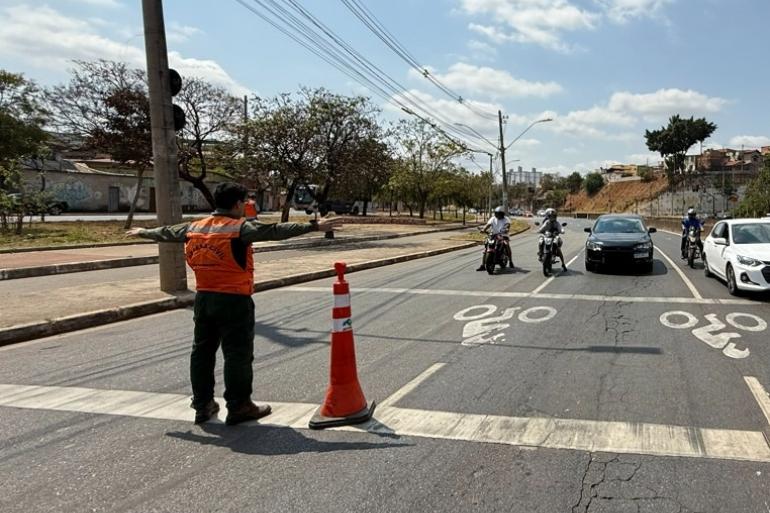 Defesa Civil realiza 6º simulado de fechamento de via na Avenida Bernardo Vasconcelos Imagem mostra um agente da Defesa Civil com colete laranja orientando o trânsito. Na foto há três motoqueiros parados e dois carros. Há árvores do lado esquerdo.