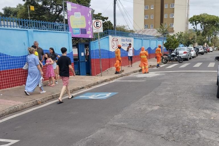 Imagem a entrada de uma escola. Em frente ao portão há várias pessoas, adultos, crianças e garis da SLU uniformizados. Há vários carros estacionados.
