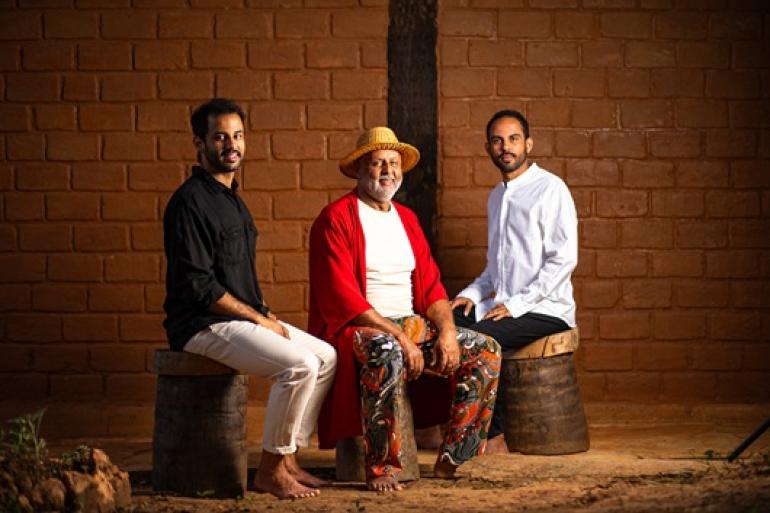 Show “Som de Casa” reúne pai e filhos em encontro musical no Teatro Raul Belém Machado A fotografia mostra Wilson Dias, Wallace Gomes e Pedro Gomes sentados em bancos de madeira diante de uma parede de tijolos de adobe. O homem do meio, mais velho, usa chapéu de palha, túnica vermelha e calça estampada; os dois homens ao seu lado usam camisas de manga longa (preta à esquerda, branca à direita).