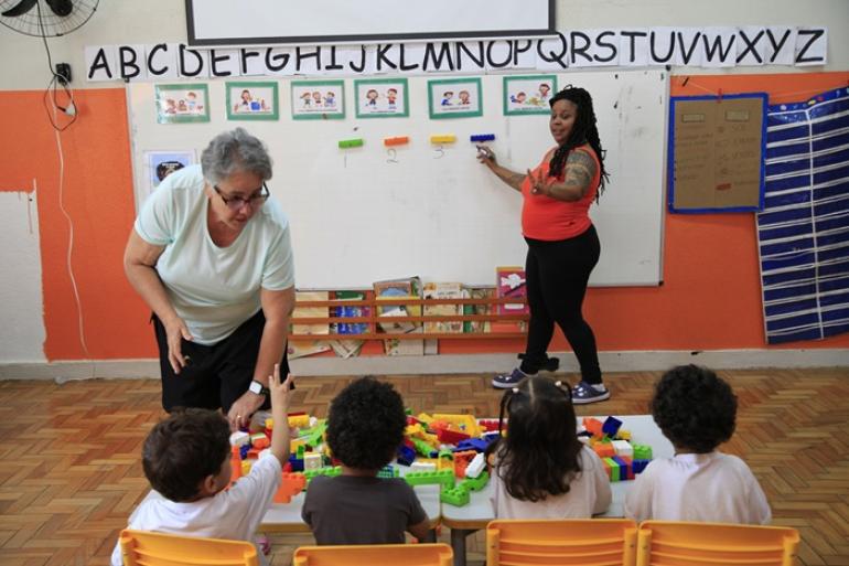 Histórias que conectam gerações na Rede Municipal de Educação marcam o Dia dos Professores em BH Duas professoras, uma de blusa vermelha e outra de blusa verde-clara, auxiliam quatro crianças em uma atividade com blocos coloridos sobre uma mesa, em uma sala de aula da Escola Municipal Marconi.