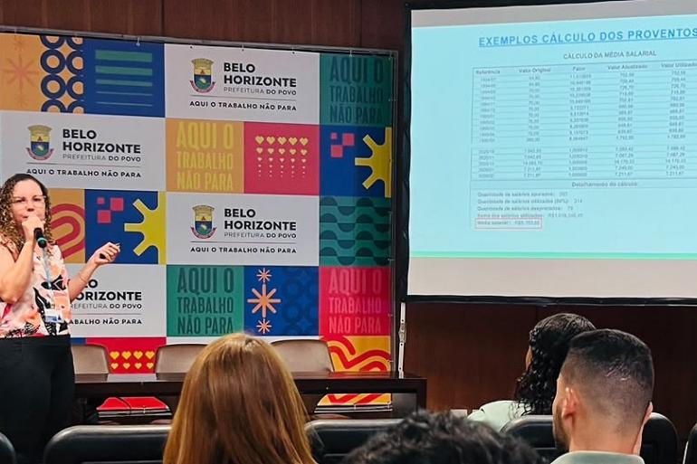 Capacitação busca aprimorar gestão de benefícios previdenciários em Belo Horizonte Registro de uma capacitação em um auditório. Uma mulher está à frente do público, falando ao microfone e apresentando o conteúdo. Ao lado dela, um telão exibe uma tabela com exemplos de cálculo de proventos. Ao fundo, há um painel colorido com a identidade visual de Belo Horizonte. Os participantes acompanham a apresentação sentados nas cadeiras do auditório.