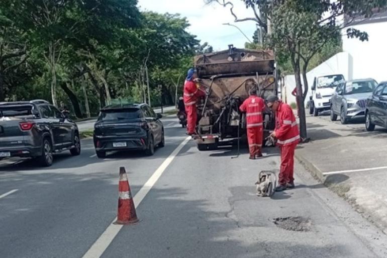 A iamgem mostra três homens com uniformes vermelhos em obras de tapa-buracos. Um deles está em um caminhão tanque e outros dois no asfalto. Ao lado do caminhão há dois carros supostamente em transito e do lado direito outros três supostamente estacionados.