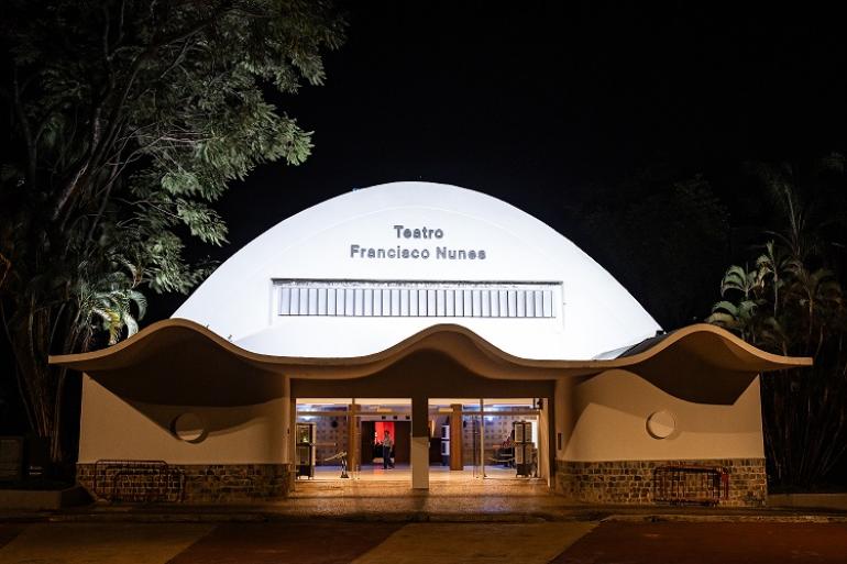 Teatro Francisco Nunes recebe espetáculo de dança inspirado em tradições mineiras Fotografia noturna da fachada do Teatro Francisco Nunes. A edificação branca e iluminada se destaca contra o fundo escuro do céu e das árvores ao redor. O prédio possui uma grande cobertura abobadada e, na entrada frontal, uma marquise com ondulações modernas sobre as portas de vidro iluminadas. No topo da fachada branca, lê-se o nome "Teatro Francisco Nunes" em letras escuras.
