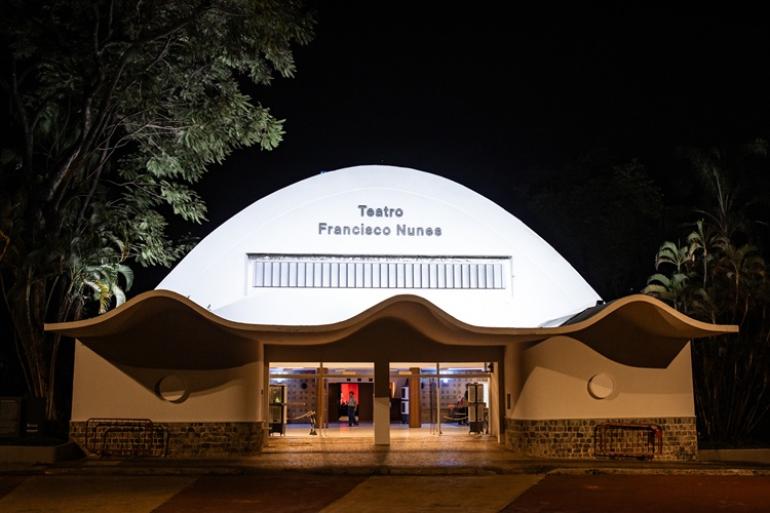Fotografia noturna da fachada iluminada do Teatro Francisco Nunes. O edifício branco possui um teto curvado em cúpula e uma pala ondulada sobre a entrada com portas de vidro, estando rodeado por árvores.