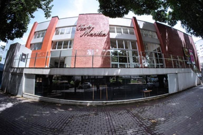 Comédia leva identificação e reflexão ao palco do Teatro Marília Fotografia de grande angular da fachada do Teatro Marília. O prédio moderno tem painéis terracota e janelas de vidro. O nome do teatro está escrito em letras brancas na parte superior central. Uma calçada de paralelepípedos e árvores estão em primeiro plano.