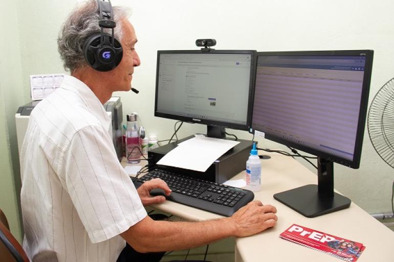 Teleconsulta amplia e agiliza o acesso à Profilaxia Pré-Exposição contra o HIV Na imagem um homem de cabelos brancos, com fones no ouvido, vestido com uma camisa branca de magas curtas está em frente a duas telas de computadores.