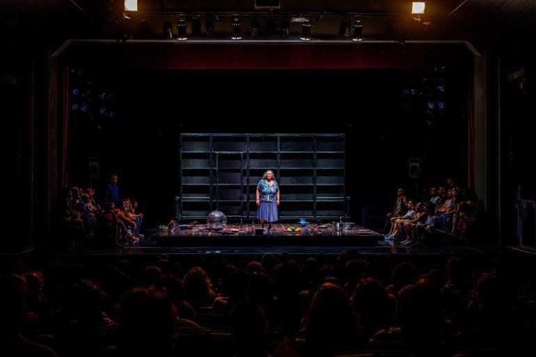 Foto da atriz Teuda Bara no palco. Ela veste uma saia azul e blusa colorida de fundo azul e possui cabelos médios, grisalhos. Espectadores assistem a atriz em cadeiras em frente ao palco e nas laterais. 