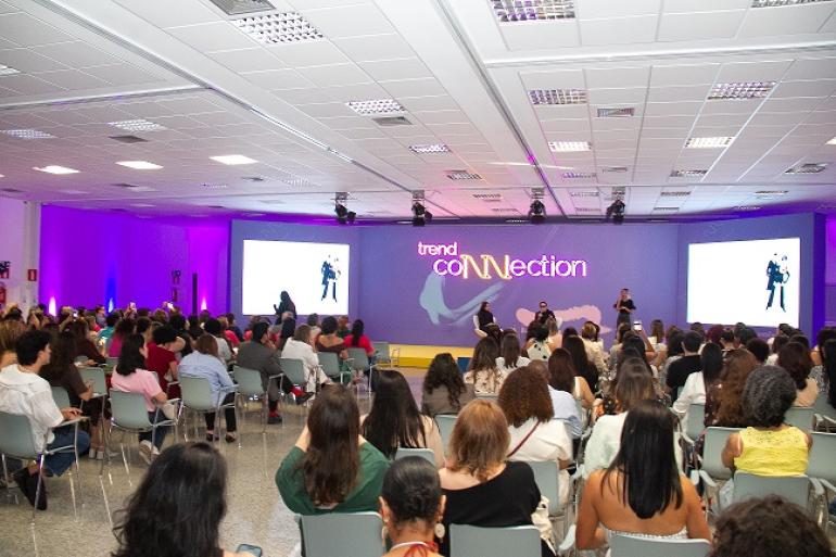 Trend Connection, evento que valoriza a indústria da moda, tem apoio da PBH Imagem mostra várias pessoas em um auditório assistindo a uma palestra. Há três pessoas no palco, duas mulheres e um homem. Uma das mulheres está de pé com um microfone na mão. No fundo há três telas roxas. na tela central há uma arte com o nome do evento.