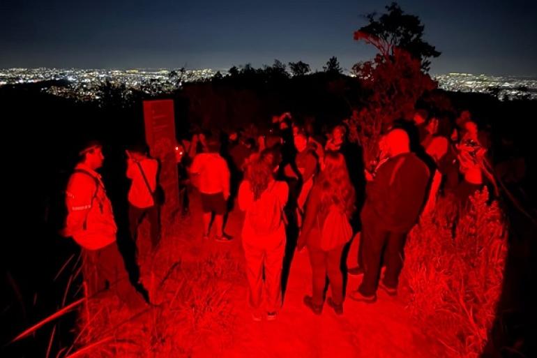 Inscrições para mais uma edição da “Trilha ao Luar” no Parque da Serra do Curral serão abertas nesta segunda-feira (21) Imagem mostra um grupo de pessoas no escuro observando a cidade iluminada ao fundo.