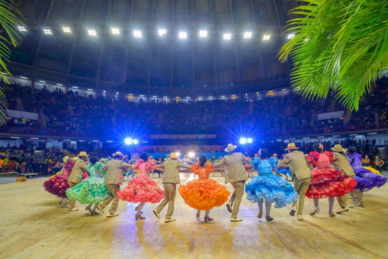 Imagem mostra bailarinos vestidos com roupas típicas de quadrilha em uma roda, debaixo de várias luzes, com o público ao fundo.