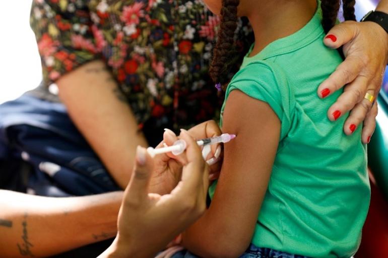 PBH aplica quase 15 mil doses de imunizantes na primeira semana da Campanha de Multivacinação Imagem mostra uma criança de blusa verde amaparada por uma mulher de blusa estampada. A criança está sendo vacinada.