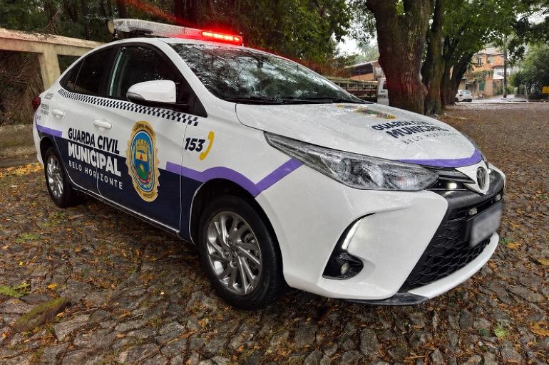 viatura da Guarda Civil Municipal de Belo Horizonte, de cor branca com faixas lilás, com o giroflex vermelho aceso no teto. O veículo está estacionado, tendo ao fundo uma área arborizada.