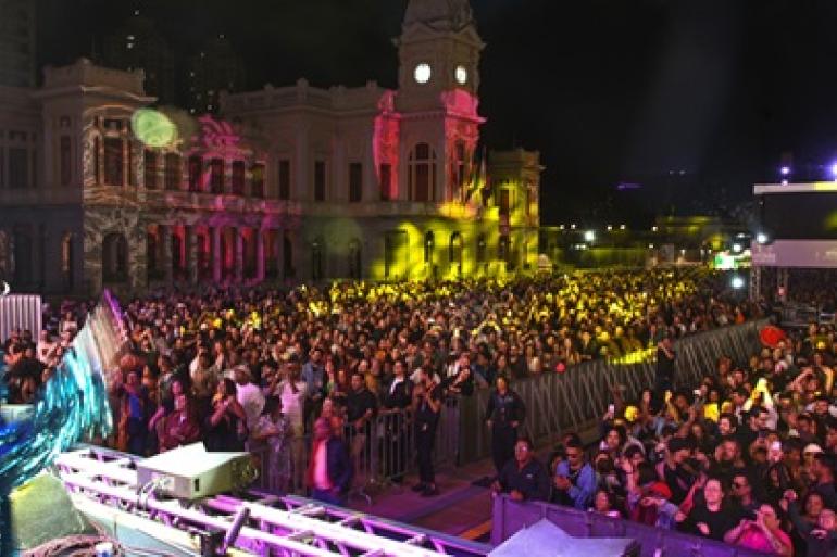Imagem noturna capturada do alto de um palco na Praça da Estação, durante um evento da Virada Cultural. Em primeiro plano, veem-se as treliças metálicas do palco e grades de proteção que separam a área de apresentação de uma multidão densa e entusiasmada, iluminada por feixes vibrantes de luzes rosa, amarela e verde. Ao fundo, destaca-se o prédio histórico da antiga Estação Ferroviária.