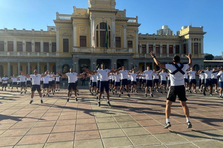 Treinão neste sábado (15) prepara os participantes para a 2ª Corrida da Guarda Municipal Fotografia ao ar livre, em dia ensolarado, mostrando um grande grupo de pessoas realizando exercícios físicos na praça da Estação.Todos estão vestindo uniforme esportivo composto por camisetas brancas de manga curta e calções escuros. De costas para a câmera, um instrutor salta com braços e pernas abertos conduzindo o exercício de polichinelo, enquanto os participantes estão posicionados em fileiras organizadas ao fundo acompanhando o mesmo movimento.