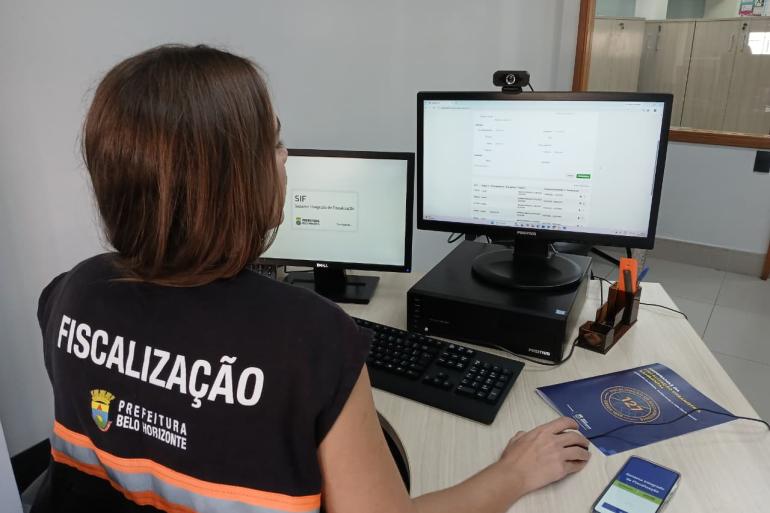 Mulher que atua na Fiscalização da PBH, usando colete, trabalha no computador