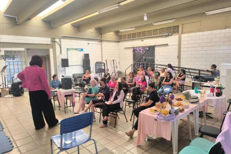 Providência em Ação amplia orientações sobre prevenção à violência contra as mulheres no Agosto Lilás Imagem de uma sala com palestra para um grupo de mulheres. À esquerda, a palestrante veste blazer rosa e calça preta, vista de costas. No centro, cerca de 15 a 20 mulheres estão sentadas em cadeiras assistindo à palestra. À direita, há uma mesa com toalha rosa servida com comidas e bebidas.