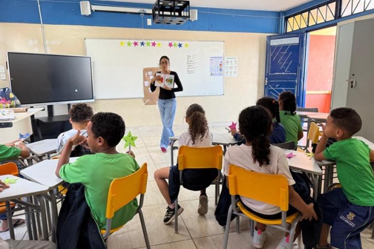 Imagem de uma professora, no fundo, mostrando um livro para uma turma de crianças uniformizadas, de costas, em uma sala de aula com paredes brancas e azuis, cadeiras amarelas e mesas brancas.