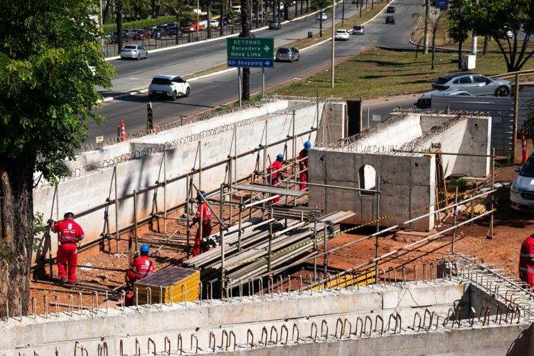 Operários vestidos de uniforme vermelho trabalham no canteiro de obras do Trevo do Belvedere, com a rodovia ao fundo