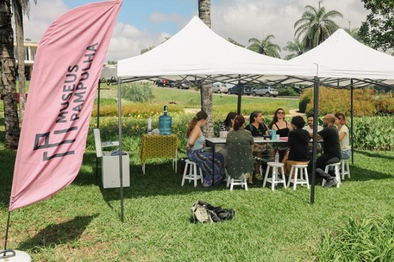 Projeto Museus Pampulha reúne arte, natureza e inclusão ao longo do mês de setembro Fotografia mostra uma atividade do projeto Museus Pampulha em uma área verde, durante o dia. Sob duas tendas brancas, cerca de dez pessoas estão sentadas ao redor de uma mesa, participando de uma atividade coletiva. À esquerda, uma grande bandeira rosa traz a logomarca do projeto. Ao redor, há gramado, jardins, palmeiras e árvores. Ao fundo, aparecem carros estacionados e parte do Museu de Arte da Pampulha.