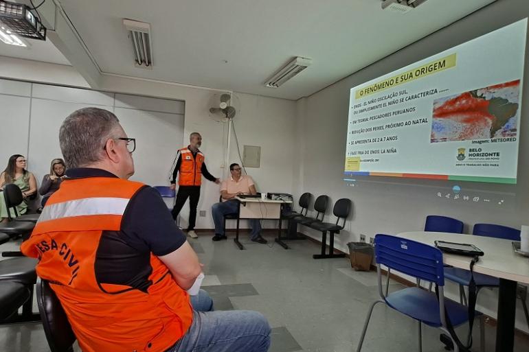 Defesa Civil inicia capacitação nas regionais para período chuvoso 2026/2027 Imagem mostra homens com coletes da Defesa Svil assistindo a uma apresentação em power point.