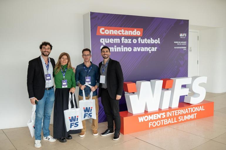 Belotur participa de evento preparatório para receber jogos da Copa do Mundo Feminina de 2027 Três homens e uma mulher, todos representantes da Belotur, estão ao ldo de um estande da Women’s International Football Summit (Wifs). Todos carregam sacolas alusivas ao evento. A mulher está de blusa verde, dois homens estão de blaser escuro e o outro de camisa azul.